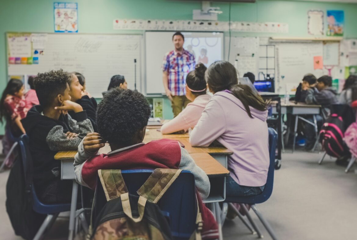 Several students in a classroom facing their teacher. The image is taken from the backs of the students who are all facing the teacher at the front of the room.
