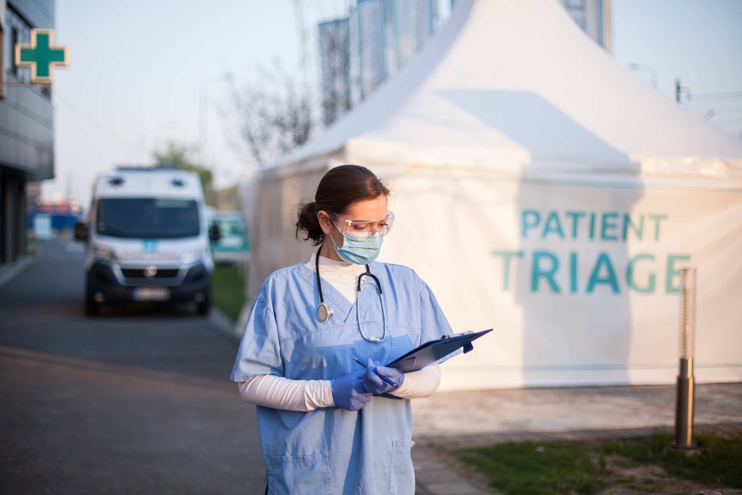 A medical professional in scrubs, mask, and protective eyewear reviewing a patient chart in front of a temporary 'Patient Triage' tent. The background features a mobile medical unit and ambulance, emphasizing the need for Temp-Air climate control and HEPA air filtration systems to maintain safe air quality in temporary healthcare environments.