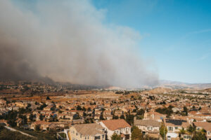 An aerial view of a suburban neighborhood with construction sites and rows of closely packed houses in the foreground, under a clear blue sky. In the background, a massive plume of thick, dark grey smoke from a wildfire rises from the hills, spreading across the horizon and partially obscuring the landscape. Construction sites and open land sit between the residential area and the fire.