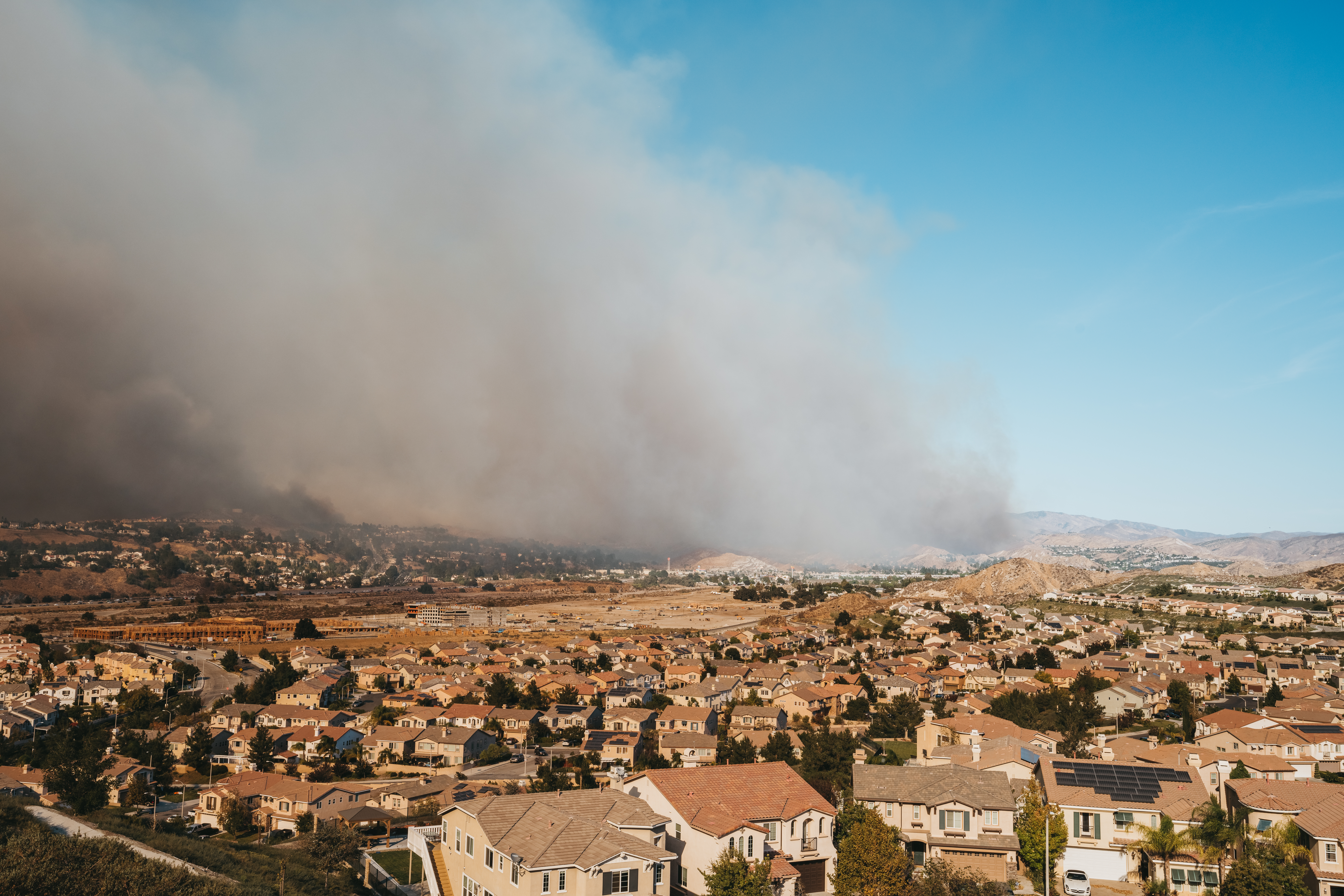 An aerial view of a suburban neighborhood with construction sites and rows of closely packed houses in the foreground, under a clear blue sky. In the background, a massive plume of thick, dark grey smoke from a wildfire rises from the hills, spreading across the horizon and partially obscuring the landscape. Construction sites and open land sit between the residential area and the fire.
