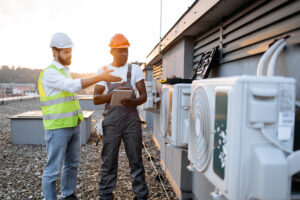 Two construction workers wearing hard hats and safety vests inspecting a large industrial commercial HVAC unit outside a facility.