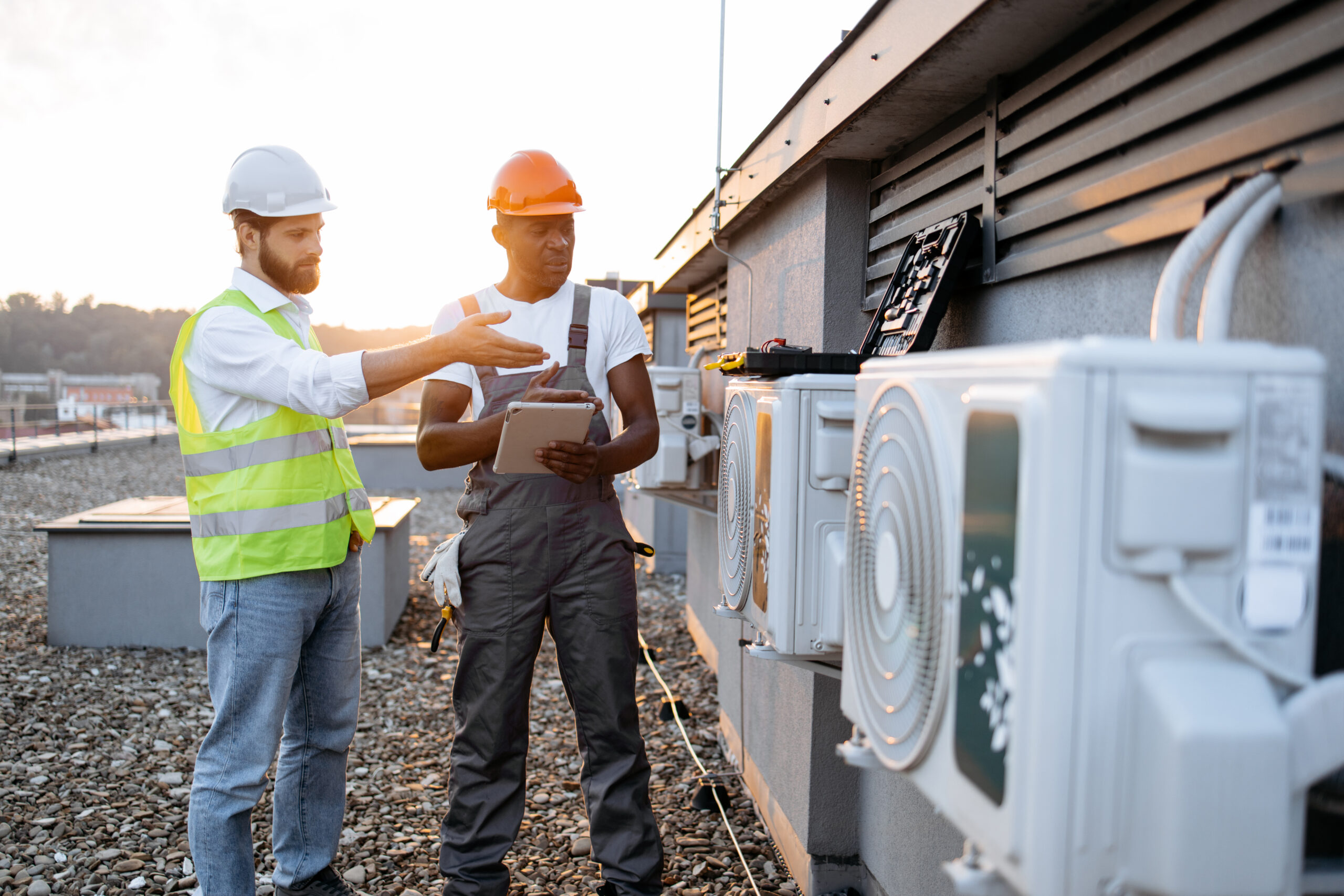 Two construction workers wearing hard hats and safety vests inspecting a large industrial commercial HVAC unit outside a facility.