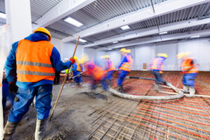 Construction workers finishing a winter concrete floor pour inside a building frame, using an industrial portable heater setup to ensure proper curing temperatures.